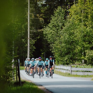 A group of cyclists riding on a rural road through a green, forested landscape. | © sportingWOMEN