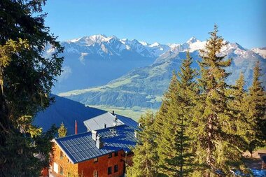 View of a mountain hut surrounded by pine trees with a stunning mountain range in the background. | © Pinzgauerhütte