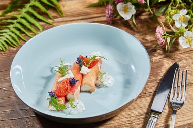 Elegant dish with fish, fruits, and edible flowers on a blue plate, placed on a wooden table with cutlery and flowers in the background. | © Nikolaus Faistauer Photography