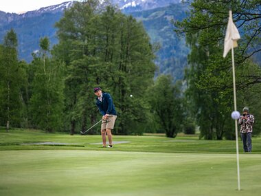 Golf player putting on a sunny green course surrounded by lush trees with mountains in the background.