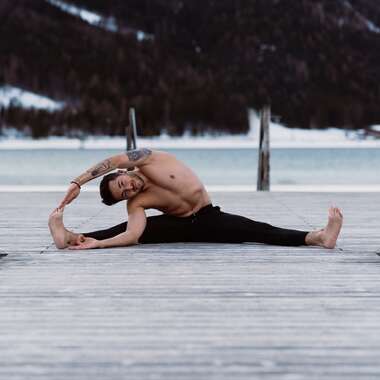 Person practicing yoga on a wooden platform in the mountains with water and snow in the background, stretching pose. | © Katharina Eder Arts 