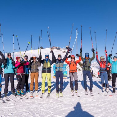 Group participating in a winter camp on snowy mountain with skis and ski poles, mountain peaks in the background, sunny day. | © Marc Stickler Photography