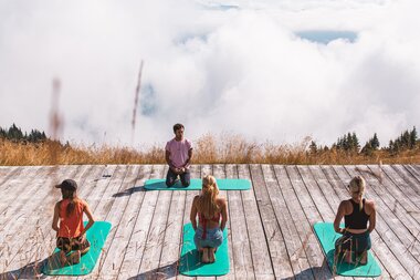 A group practicing yoga on a platform in the mountains, overlooking clouds and sky. | © Johannes Radlwimmer 
