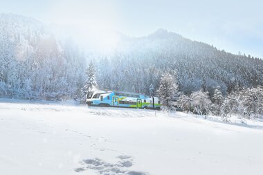 A train travels through a snowy winter landscape with trees and mountains in the background. | © Westbahn