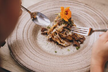Person eating a dish with meat and shrimp, garnished with an edible flower on a round, textured plate. | © Zell am See-Kaprun Tourismus