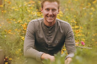 A person sitting in a blooming meadow with colorful flowers, smiling at the camera. | © Zell am See-Kaprun Tourismus