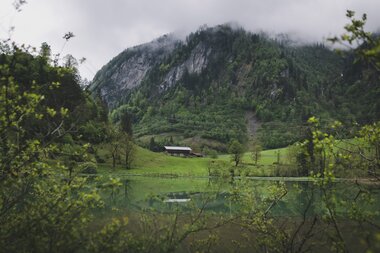 The Sigmund Thun Gorge near Kaprun in May, surrounded by mountains, with calm water in the foreground and cloudy sky. | © Oberhauser Photography