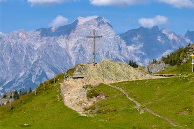 Mountain cross on a green meadow with mountain peaks in the background under a blue sky. | © Zell am See-Kaprun Tourismus