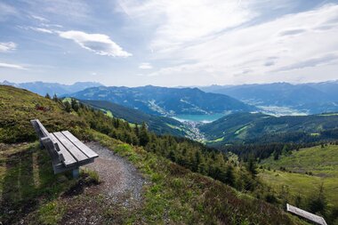 Viewpoint on a high plateau overlooking mountains, valleys, and a lake in the background, with a bench along the path. | © Zell am See-Kaprun