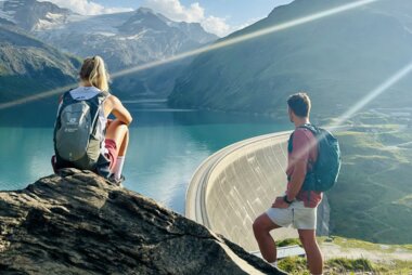 Two hikers with backpacks by the edge of a large high mountain reservoir surrounded by mountains on a clear day. | © Zell am See-Kaprun