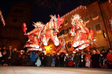 Dancing figures in colorful costumes at a festive event in a city at night. | © Nikolaus Faistauer Photography