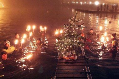 People swimming in water at a festive Christmas event with lights, a Christmas tree, and a celebratory atmosphere. | © Schweinoster