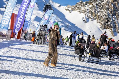 Person at a winter event in a snowy ski resort area, surrounded by ski lifts, tents, and other people. | © Tim Mengerink