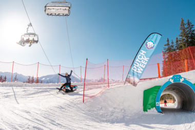 Skier on a snowy slope during a winter event, sunlight, red safety fences, and a snowboard with a tube passing through a tunnel. | © Dutchweek Winter