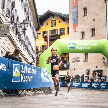 Participants running in the IRONKIDS event in Zell am See-Kaprun, on a city street with banners and a green finish arch. | © Zell am See-Kaprun, JOE
