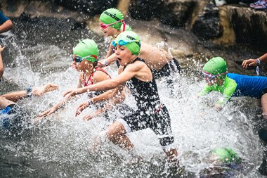 Children participating in the IRONKIDS race in water, wearing swimming caps and goggles, splashing water. | © Zell am See-Kaprun, JOE