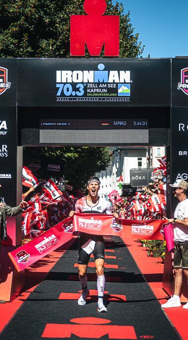 Winner crossing the finish line at IRONMAN 70.3 Zell am See-Kaprun 2025, surrounded by cheering fans and organizers. AI-generated text. | © Zell am See-Kaprun, JOE