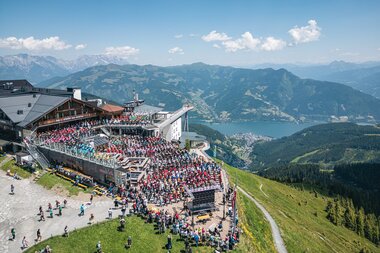 Large outdoor event on a mountain terrace with views of mountains and a lake, many visitors, and an outdoor stage. | © Zell am See-Kaprun, JOE