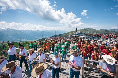 Large line dance event with many participants in traditional costumes and hats against an alpine mountain backdrop on a sunny day. | © Zell am See-Kaprun, JOE