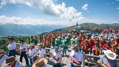 Large line dance event with many participants in traditional costumes and hats against an alpine mountain backdrop on a sunny day. | © Zell am See-Kaprun, JOE