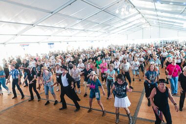 Large group of people practicing line dance in a festival tent with bright lighting. | © Zell am See-Kaprun, JOE