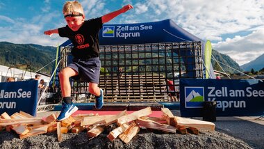 Child splitting wood during a kids' marathon in Zell am See-Kaprun with mountain scenery in the background. | © Zell am See-Kaprun Tourismus