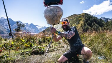 Participant in a Spartan Race holding a large weight on the outdoor course with mountains in the background. | © Zell am See-Kaprun Tourismus