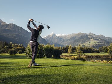 Person playing golf on a green field with mountains and a lake in the background on a sunny day. | © Zell am See-Kaprun Tourismus