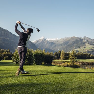 Person playing golf on a green field with mountains and a lake in the background on a sunny day. | © Zell am See-Kaprun Tourismus