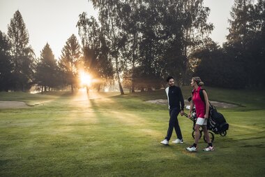 Two people playing golf on a well-maintained golf course at sunrise, with trees in the background. | © Zell am See-Kaprun Tourismus