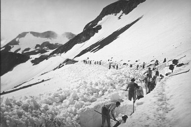 People clearing snow on the Großglockner High Alpine Road, using shovels in a snowy mountain landscape. | © Großglockner Hochalpenstraße 