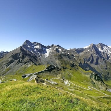 View of alpine landscape with winding road and tall mountains, snow on peaks, under a clear blue sky. | © Großglockner Hochalpenstraße 