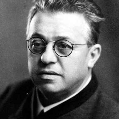 Black and white photo of a person with round glasses, short hair, and formal attire.