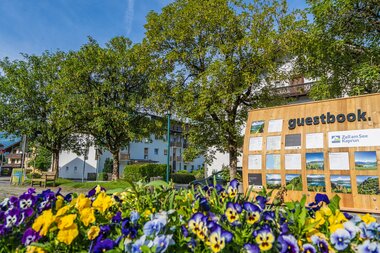 Flowers in the foreground, large trees, and a guestbook display nearby, with buildings under a blue sky in the background.