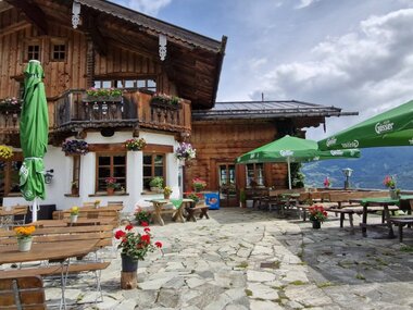 A mountain restaurant with an outdoor seating area, tables, benches, and umbrellas, surrounded by flowers and mountains. | © Schoberalm