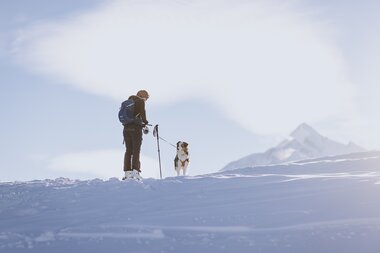 Hiker with backpack ski touring with a dog in snowy mountain landscape, sky with few clouds in the background. | © JFK Photography