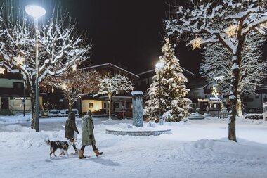 Two people walk with a dog through a snow-covered, festively illuminated town in winter. | © JFK Photography