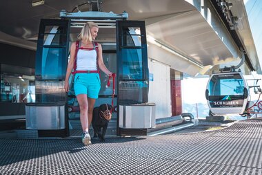 Person disembarking a cable car with a dog at a mountain station, mountain scenery in the background. | © Zell am See-Kaprun Tourismus