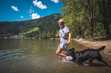 Person playing with a black dog at the lakeshore in a mountainous landscape, during summer on a sunny day. | © Zell am See-Kaprun Tourismus