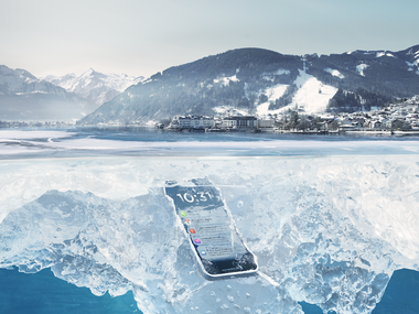 A smartphone is embedded in an ice layer on a frozen lake, surrounded by snow-covered mountains and a winter landscape background. | © Zell am See-Kaprun