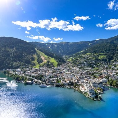 Aerial view of Zell am See with lake, town, and surrounding mountains on a sunny day. AI-generated text. | © Nikolaus Faistauer