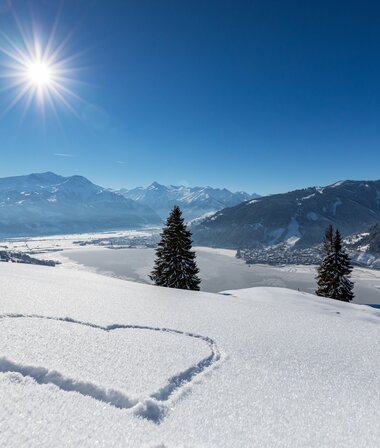 Winter landscape with snow-covered fields, trees, a heart in snow, mountains in the background, and a clear blue sky with the sun. | © Nikolaus Faistauer