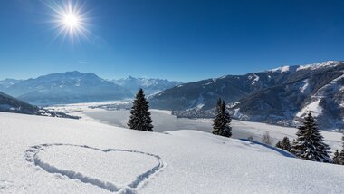 Winter landscape with snow-covered fields, trees, a heart in snow, mountains in the background, and a clear blue sky with the sun. | © Nikolaus Faistauer