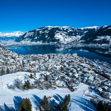 Aerial view of a snowy town by a lake surrounded by mountains in winter. | © Christian Mairitsch