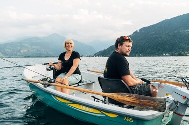 Two people sitting in a small boat on Zeller See, surrounded by mountains and water, while fishing. | © Zell am See-Kaprun Tourismus