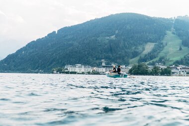 A person boating on Lake Zeller with mountains and a lakeside town in the background. | © Zell am See-Kaprun Tourismus