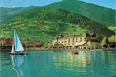 Scenic view of a calm lake with a sailboat, mountains in the background, and a building by the shore.