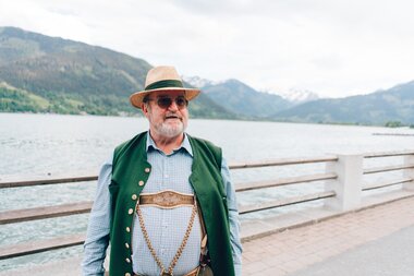 Person by a lakeside surrounded by mountains, dressed in traditional attire with a hat, glasses, and a vest. | © Zell am See-Kaprun Tourismus