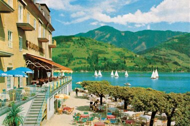 Exterior view of the Grand Hotel by a lake with terraces, tables, and sailboats on the water, surrounded by green mountains.