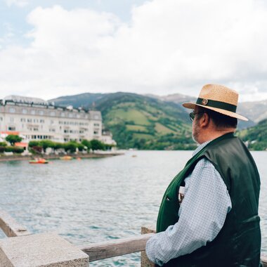A person wearing a hat and jacket looking at a lake with mountains in the background, with a stone railing in the foreground. | © Zell am See-Kaprun Tourismus